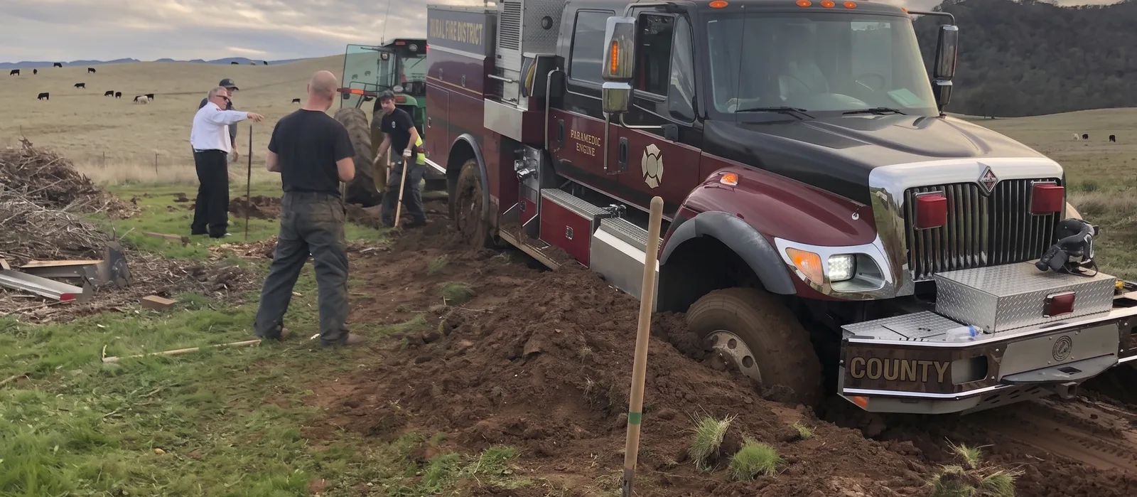 Crew working a wheels-deep fire engine recovery in soft ground with shovels and a tractor.